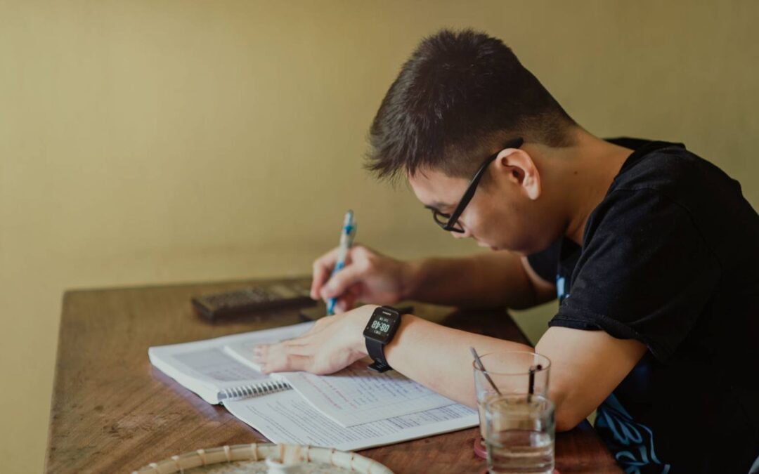 A young man concentrates on studying at his desk, taking notes indoors.